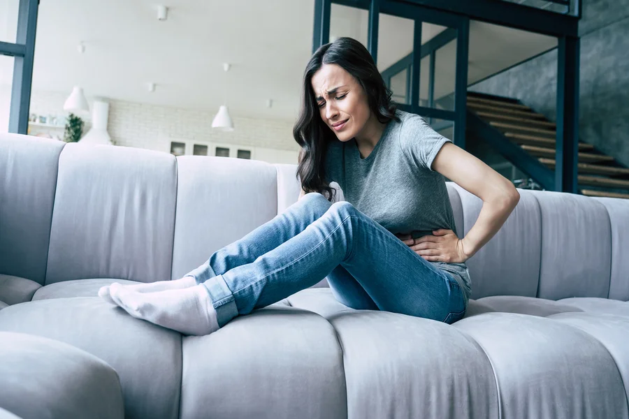 Frau mit Endometriose-Schmerzen sitzt mit schmerzverzerrtem Gesicht und angewinkelten Beinen auf Sofa und hält sich den Bauch